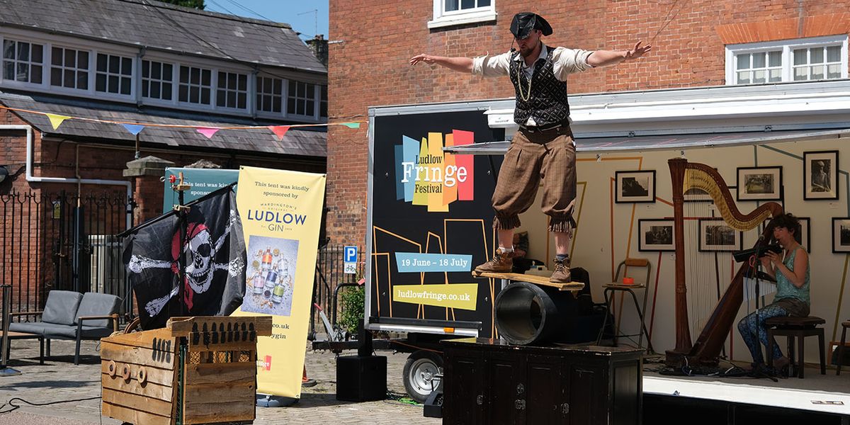 A pirate balancing on a plank on a pipe on a box, at the Ludlow Fringe, as a harp player looks on